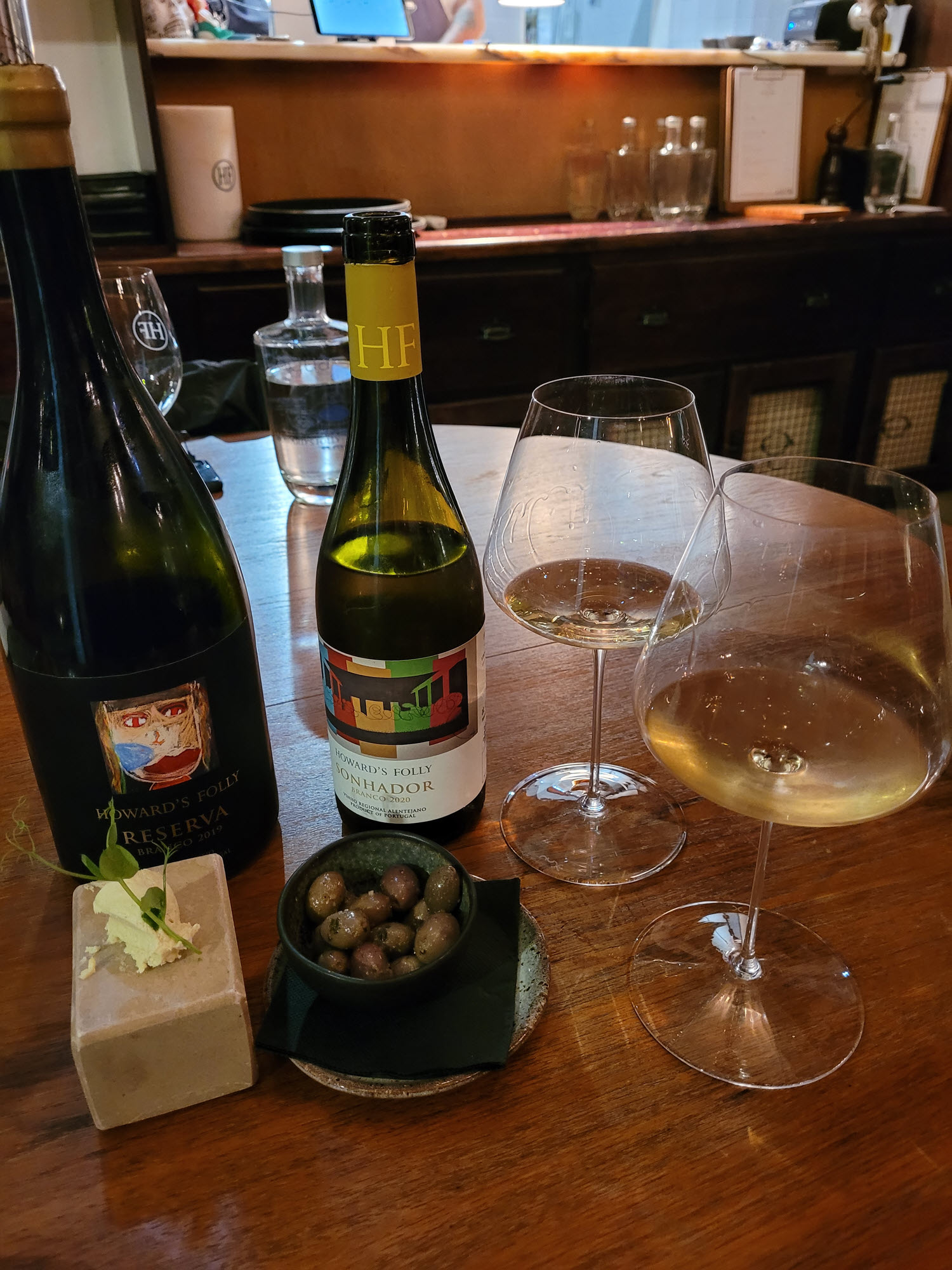 White and red wine bottles with white wine in wine glasses and a dish of olives on the side
